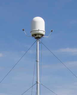 White dome radar antenna atop a tall metal mast with guy wires against a blue sky