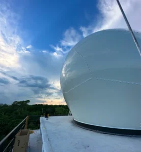 Large white domed structure on a rooftop with a metal railing, against a blue sky with clouds and distant trees in the background.
