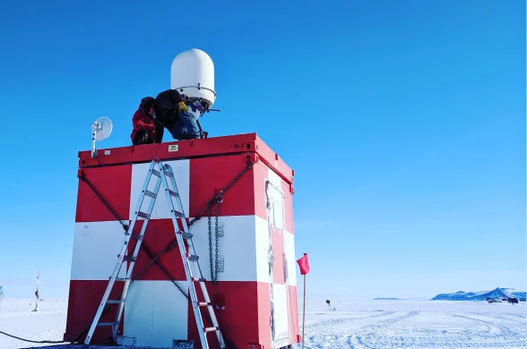 Technician on top of a red-and-white weather radar hut in a snowy landscape, ladder leaning against the hut.